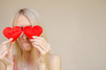 Lifestyle concept: sad, resentful attractive blonde with two small red hearts in her hands, closing her eyes with heart symbols, standing on a light background. Valentine's day