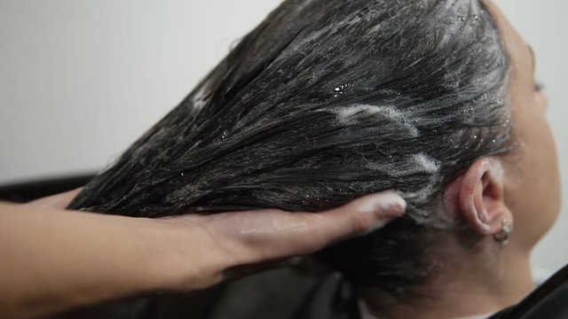 Close up footage of washing head of woman with long, dark hair with shampoo. Massaging the head sculp. Close up hands of hairdresser washing hair on head, foaming. Relaxing spa procedures