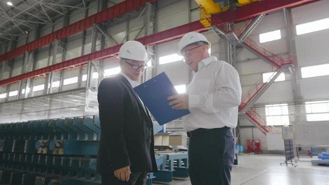 Businessman and engineer talk looking checking blueprints. Two business people or contractors discussing project with documents standing in large workshop of modern factory