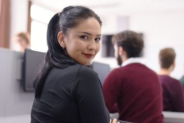 Happy young black-haired female student smiling into camera during comp lab class. Young caucasian lady student looking at camera smiling learning inside computer lab. Education concept.