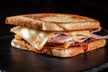 Homemade sandwich made of toast bread, cutlet, bacon, cheddar cheese, ketchup sauce. Sandwich on on black slate, on a black background.