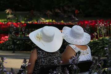 Two older women wearing wide white hats sitting together in a spa park full of colorful blooming flowers