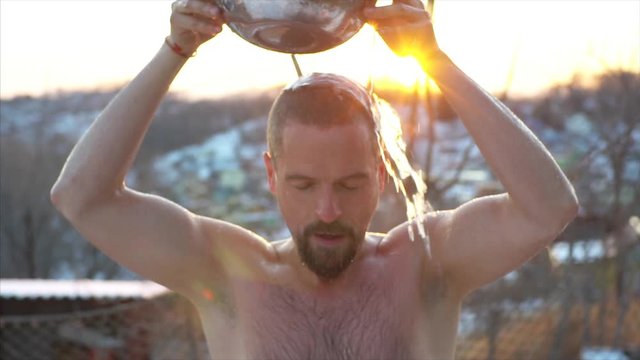 Slow motion, portrait of bearded man pouring cold water on himself from a bowl and enjoying winter freshness outdoor. Sunset