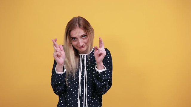 Attractive caucasian young lady holding finers crossed, make wish, waiting for special moment over isolated orange background in blue shirt with white polka dot. Lifestyle concept