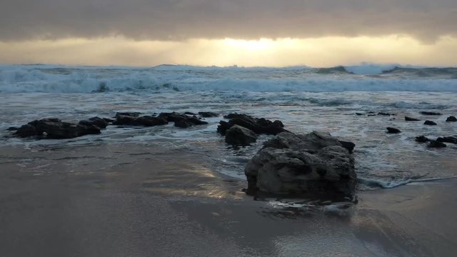 Waves gently washing and crashing across large rocks on a beach during sunrise