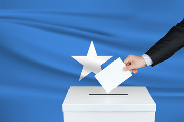 Election in Somalia. The hand of man putting his vote in the ballot box. Waved Somalia flag on background.