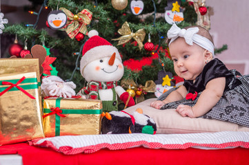 Fototapeta na wymiar A white baby in Christmas tree decoration with black dress and white bow leaning on her hand looks at happy gifts