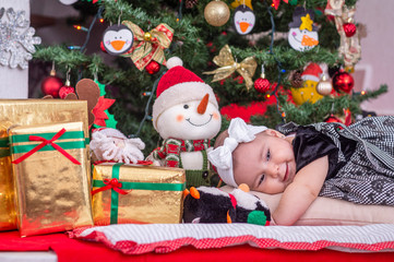 A white baby in Christmas tree decoration with black dress and white bow lying on the cushion smiles