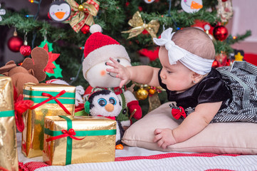 Fototapeta na wymiar A white baby in Christmas tree decoration with black dress and white bow tries to reach doll