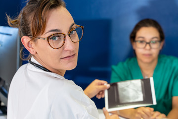 doctor with glasses and an x-ray in her hand looking at her back with a blurred nurse in the background