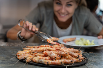 Blonde girl crouching smiling picking up a shrimp with a pair of tweezers at a self-service restaurant food counter