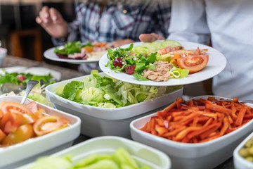 Detail of the hands of a man holding a salad plate in front of a food counter at a self-service restaurant