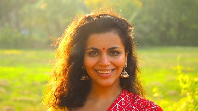Portrait of beautiful girl smiling to camera with gold earrings and red bindi tilla on forehead green field rice plantation background