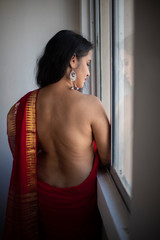 Fototapeta na wymiar Back portrait of young and attractive Indian Bengali brunette woman in traditional red sari standing in seductive way in front of glass window in white background. Indian lifestyle and bold fashion.