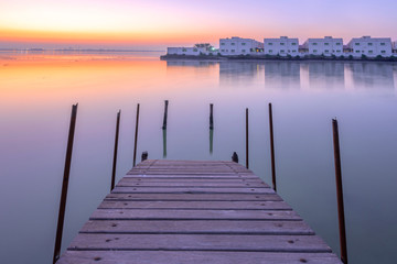 Wooden bridge out on the water over sunrise colorful sky with traditional local house in the island background, Bahrain.