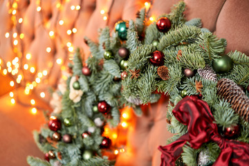Closeup Beautiful festive wreath of fresh spruce on sofa. Christmas mood. Xmas tree. Bokeh of Garland lights on background
