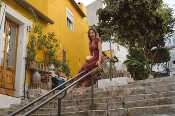 Young traveling woman wearing long red dress walking on Lisbon streets looking at beautiful view, have fan
