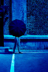 Walking woman in grey sponge bags and boots with upper part of her body hidden behind black mockup umbrella. On yje background of red brick wall. Classic, blue monochrome, trend 2020