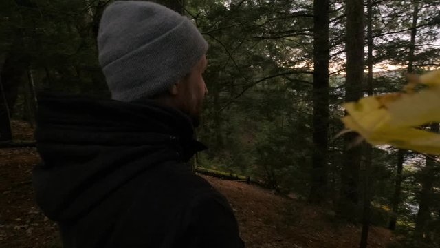 Contemplative Young White Male in Forest Watches Sunset in Slow Motion. A Person in Black Coat and Grey Hat Thinks about Life in Natural Green Trees and Wind, Handheld medium close up pan reveal