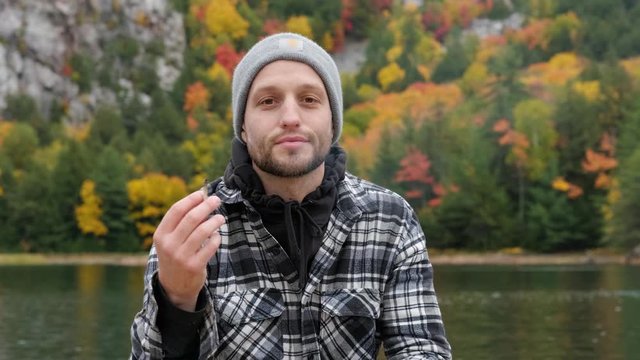 Attractive young man smiles and smokes a weed joint in slow motion, handheld. Stunning autumn foliage color with white male person enjoying cannabis in plaid shirt and hat in canoe, outdoor pan