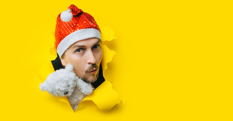 A young guy with a beard and a mustache with teddy bear in a Santa Claus or Snow Maiden hat with white pigtails peeps out of a torn hole in yellow paper. The concept of Christmas, New Year. Copy space
