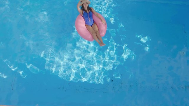 Beautiful brunette woman relaxing on inflatable pink ring in swimming pool. Aerial dolly shot