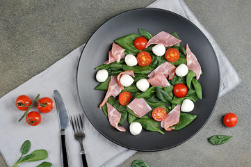 Salad with Parma ham, mozzarella cheese, cherry tomatoes and leafy spinach on a black plate.  In the frame a napkin and cutlery.  View from above.  Close-up.