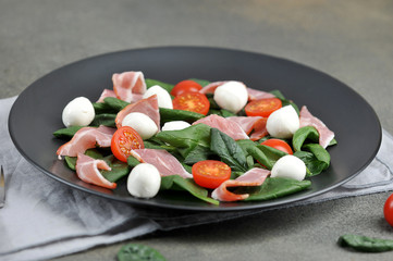 Salad with Parma ham, mozzarella cheese, cherry tomatoes and leafy spinach on a black plate. Close-up. Macro shot.