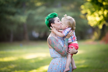 A portrait of Caucasian young woman with green hair and tattoo holding a blond toddler boy in her hands. Hugging, Mother's day concept
