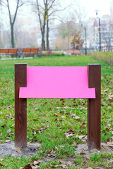 Blank pink wooden board for information and indications in the park, with a background of green grass covered by autumn leaves