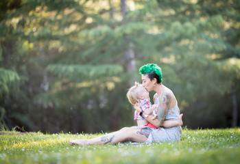 Young mother with green short hair is sitting on green grass, smiling, looking at her toddler son on her laps. Mother's day concept