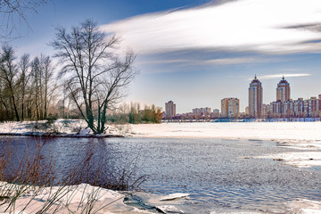 Obraz premium View of willow trees and poplars close to the frozen Dnieper River in Kiev during winter. High buildings in the background.
