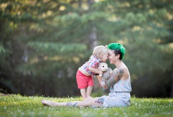 Young mother with green short hair is sitting on green grass, smiling, looking at her toddler son on her laps. Mother's day concept