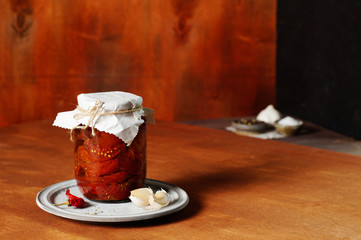 Dried, at home, tomatoes are stacked in a jar and filled with oil and spices. Standing on a ceramic plate next to the garlic and hot pepper. In the background, gravy boats with additives.Copy space.