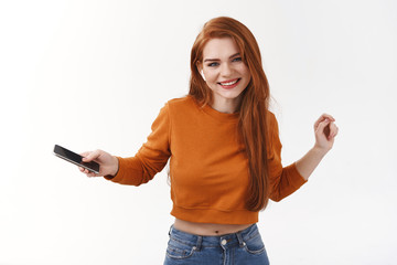 Fototapeta premium Carefree pretty caucasian redhead woman in orange cropped sweater, holding smartphone, dancing and moving rhythm music, listen catchy song in wireless earphones, smiling camera happy