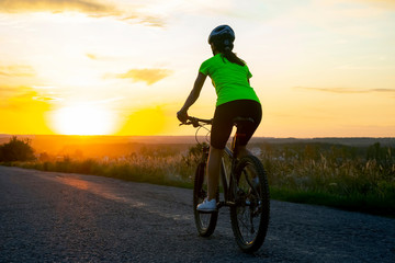 Naklejka na ściany i meble Beautiful girl cyclist rides a bicycle on the road in the sunset. Healthy lifestyle and sport. Leisure and hobbies