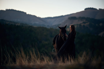 Fototapeta na wymiar Beautiful young girl in a black dress hugs her black horse at sunset in the mountains