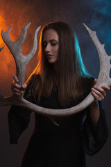 Forces of Nature craft concept. Close up portrait of young beautiful witch making ritual with deer horns over night rain and volcanic eruption background. Studio shot
