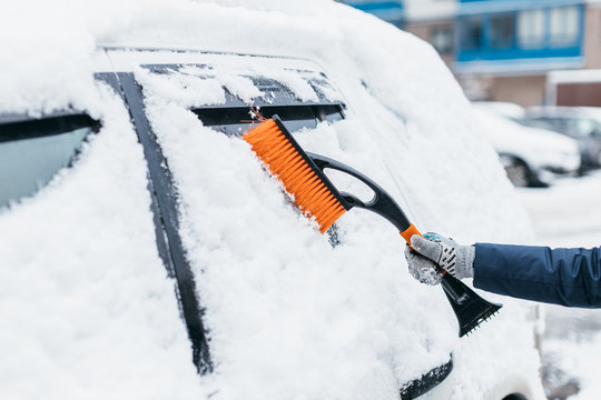 Cropped photo of woman hand in grey mitten brushing the snow from the car window. Selective focus on orange broom. Vehicle covered with snowdrifts. Heavy snowfall. Winter and seasonal concept.