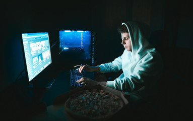 Young male gamer in white hoodie plays online games on computer and eats delivery pizza and types...