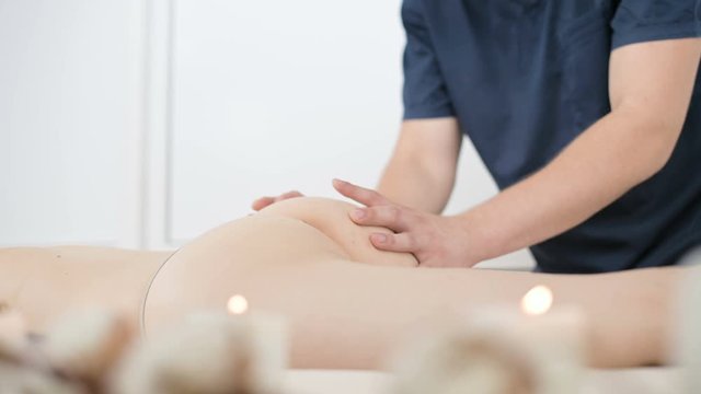 Close-up of a male physiotherapist doing a light massage to warm up the hips and buttocks of a young woman in a professional spa salon next to a sprig of cotton. 4K massage body care concept