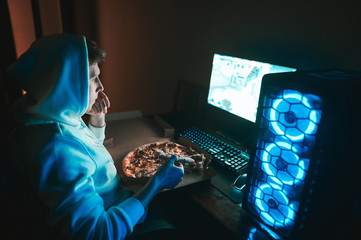 Portrait of concentrated young man eating pizza at night and looking at computer screen. Gamer...