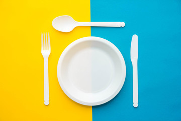 Plastic white fork, knife, spoon and plate on yellow and blue background. Cooking utensil. Top view. Minimalist Style. Copy, empty space for text