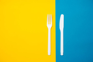 Plastic white fork and knife on yellow and blue background. Cooking utensil. Top view. Minimalist Style. Copy, empty space for text