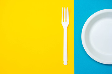 Plastic white fork and plate on yellow and blue background. Cooking utensil. Top view. Minimalist...