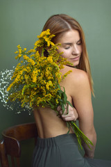 Beautiful young shirtless woman with ginger hair hugging herself the back with big flower bouquet of mimosa, sitting on wooden chair Self-care concept.