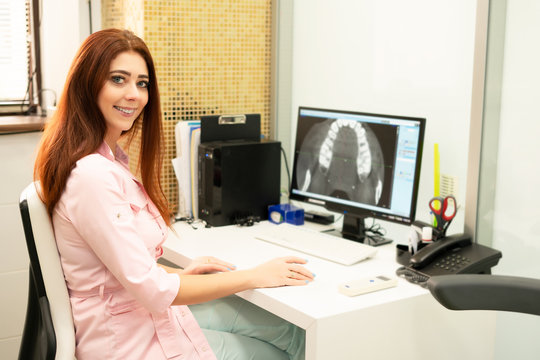 A female dentist doctor is sitting at a table, on a computer a CT scan of the jaw. The doctor is dressed in professional clothes.