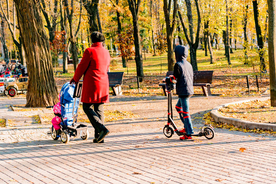 Grandmother In Red Jacket Walks With Grandchildren In The Park. One Of Them On A Stroller And Second One On A Scooter. Family Stroll. Life. Outdoor. City. Urban