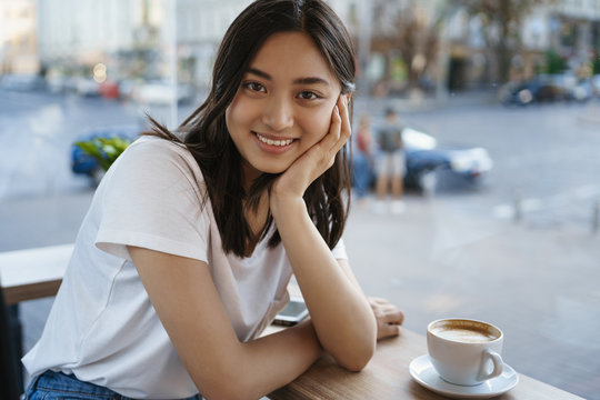 Urban lifestyle oncept. Lovely asian girl sitting coffee table, drink cappucino flirty gazing camera and smiling as listening boyfriend, talking to her date at fancy cafe, city streets on background