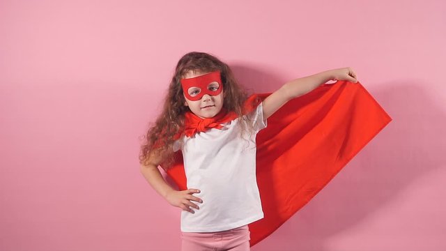 Attractive cute kid wearing red superhero suit and mask standing at isolated over pink background.